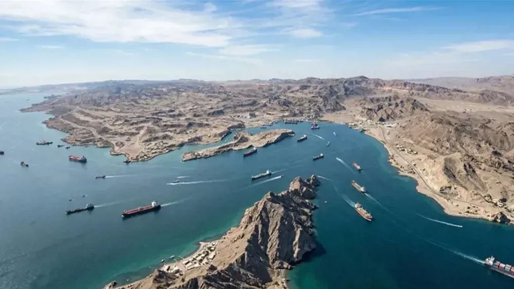 Aerial view of a deep blue harbor with numerous cargo ships along a rugged desert coastline.